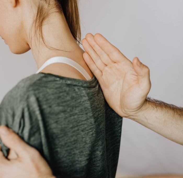 Chiropractor assessing a female patient's neck and shoulder alignment from behind, with one hand on her upper spine and the other on her shoulder.